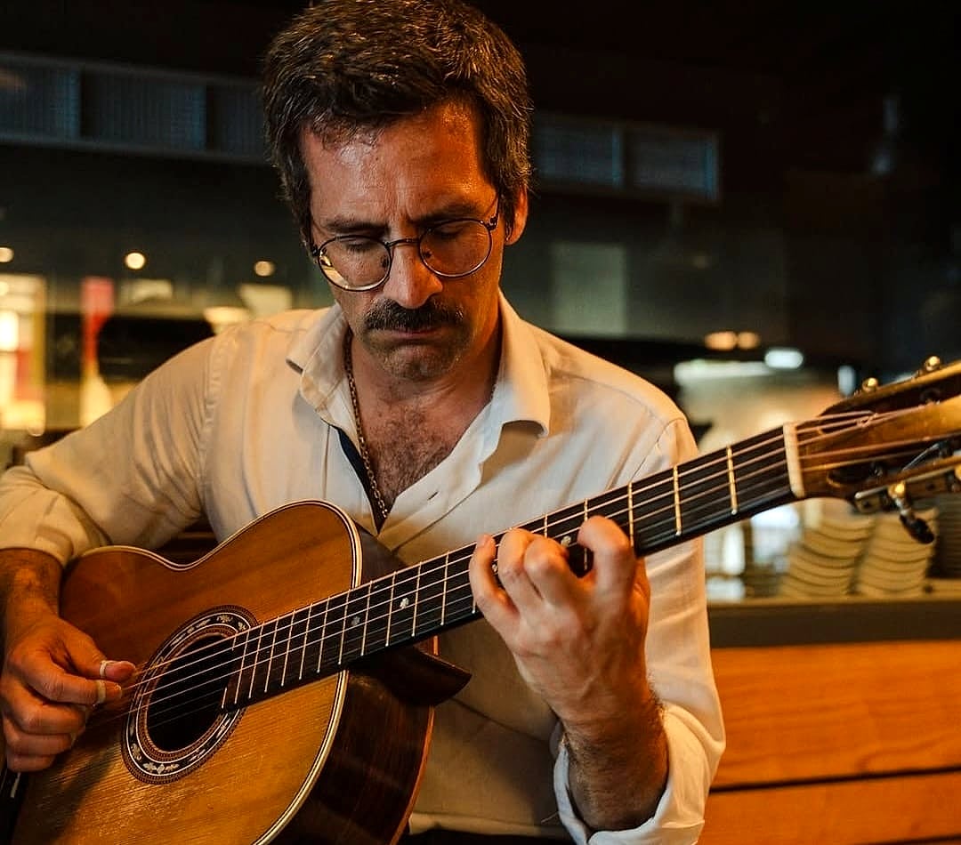Luís Roquette a tocar guitarra, de camisa branca, com o olhar baixo sobre o braço do instrumento